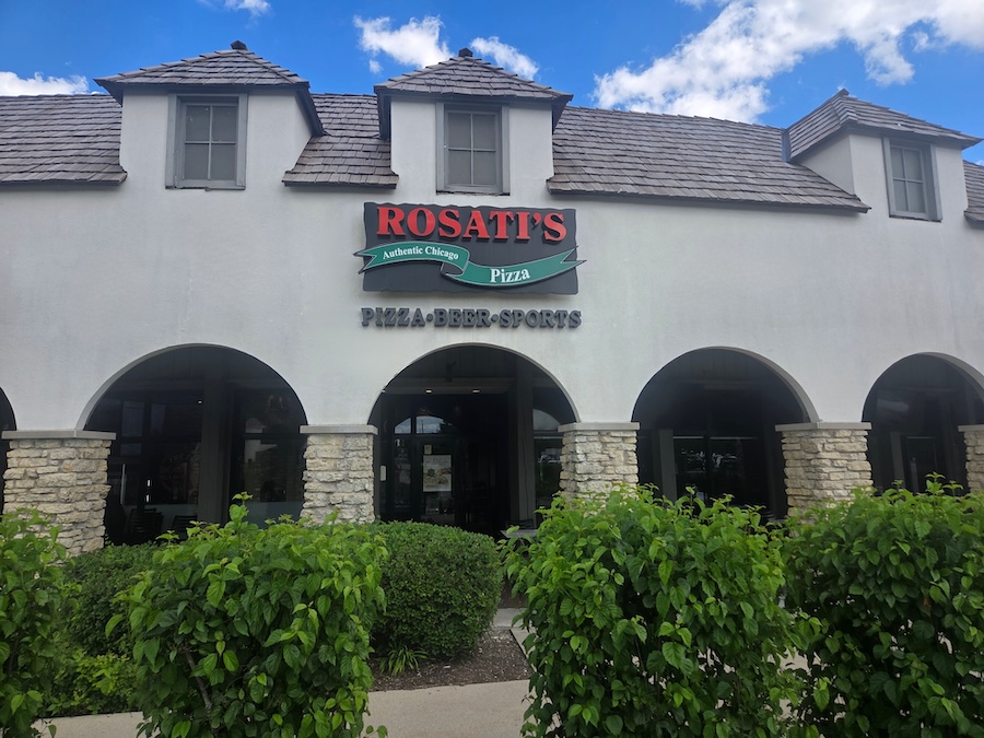 Exterior of a Rosati’s Pizza restaurant in Bannockburn, Illinois, featuring the Rosati’s sign on a white stucco building with stone columns, arched entryways, landscaped greenery, and a blue sky with clouds.