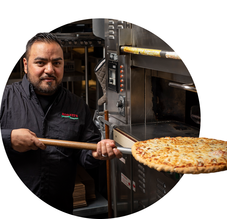 A male chef in a black shirt holds a large pizza on a wooden peel, standing next to a commercial pizza oven in a kitchen.